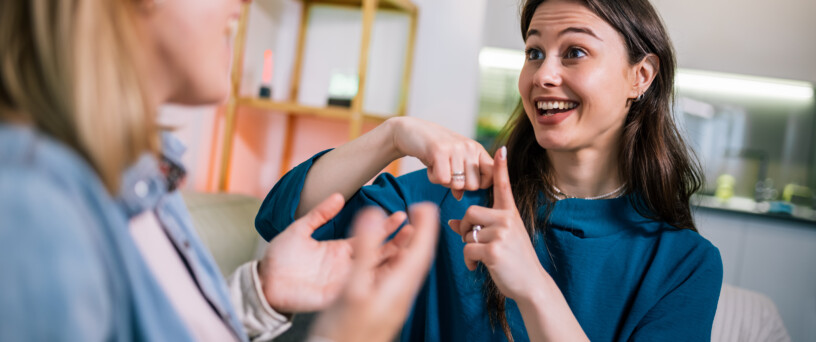 Two young girls are using sign language to talk to each other. One of them is wearing a hearing aid. They are smiling and enjoying the conversation. Mobile