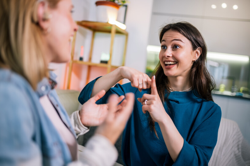 Two young girls are using sign language to talk to each other. One of them is wearing a hearing aid. They are smiling and enjoying the conversation.